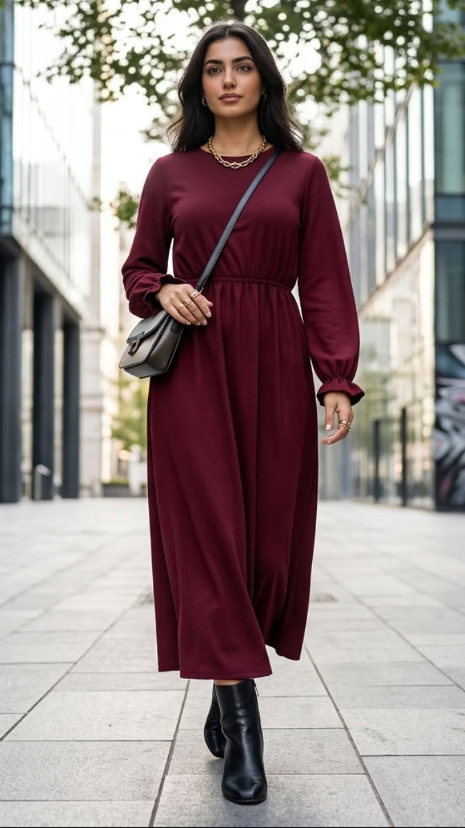 A woman walking down a modern city street wearing a long-sleeve, high-waisted maxi dress in deep burgundy. She is carrying a small gray shoulder bag and wearing black ankle boots.