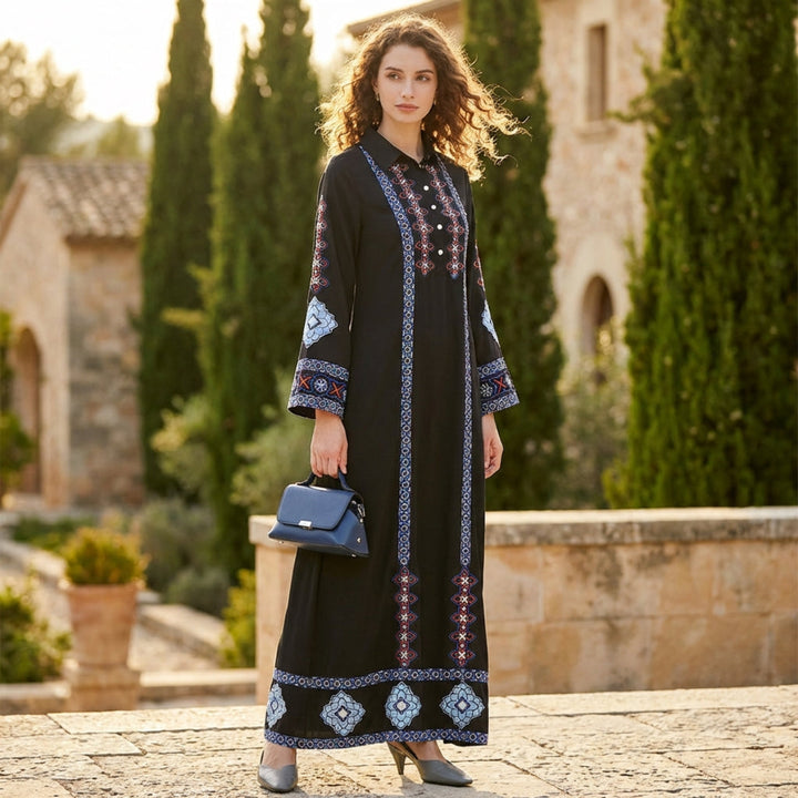 Woman in a long black dress with patterned sleeves and hem, holding a blue handbag, standing in a sunlit outdoor setting.