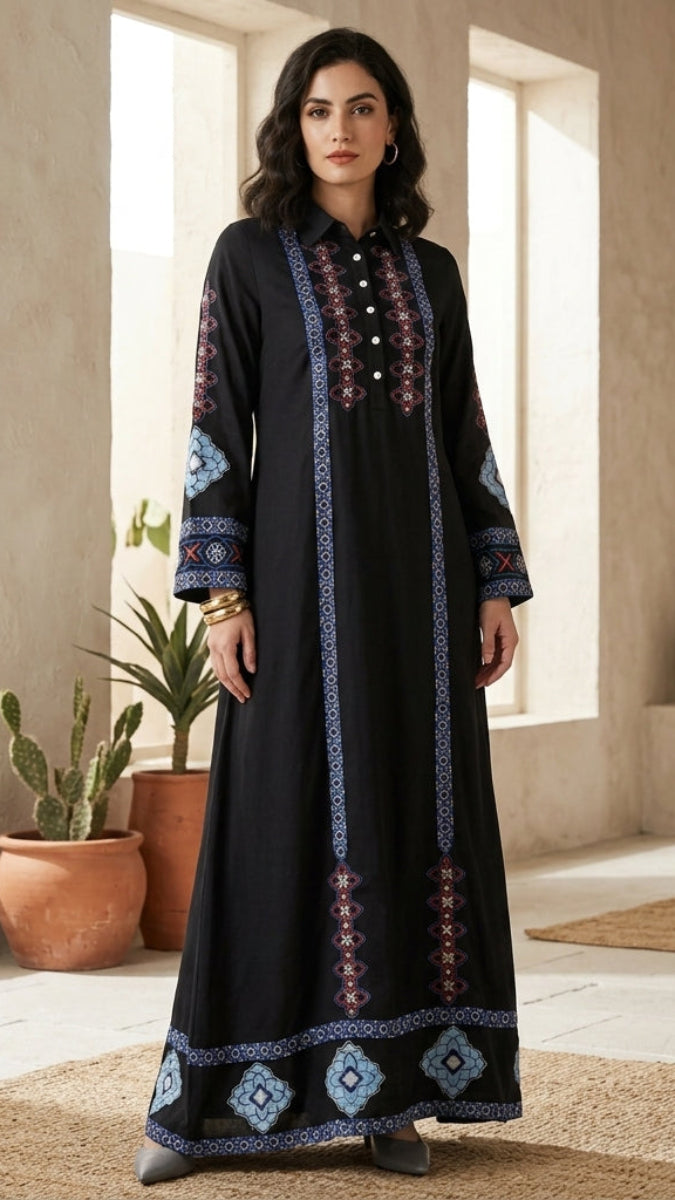 A woman models a long black maxi dress with intricate blue, red, and white geometric embroidery on the neckline, sleeves, and hem, set against a warm, sunlit interior background with potted cacti.