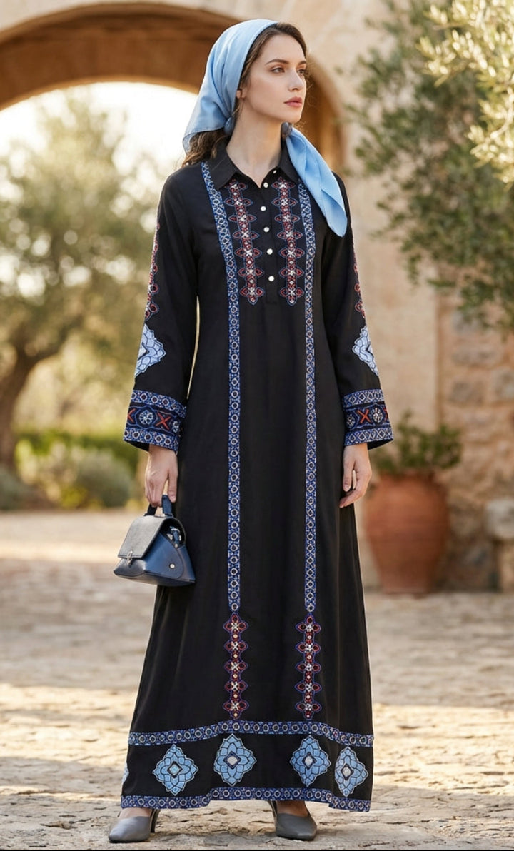 Woman in a black dress with blue embroidery standing in an outdoor setting with stone archway and trees.