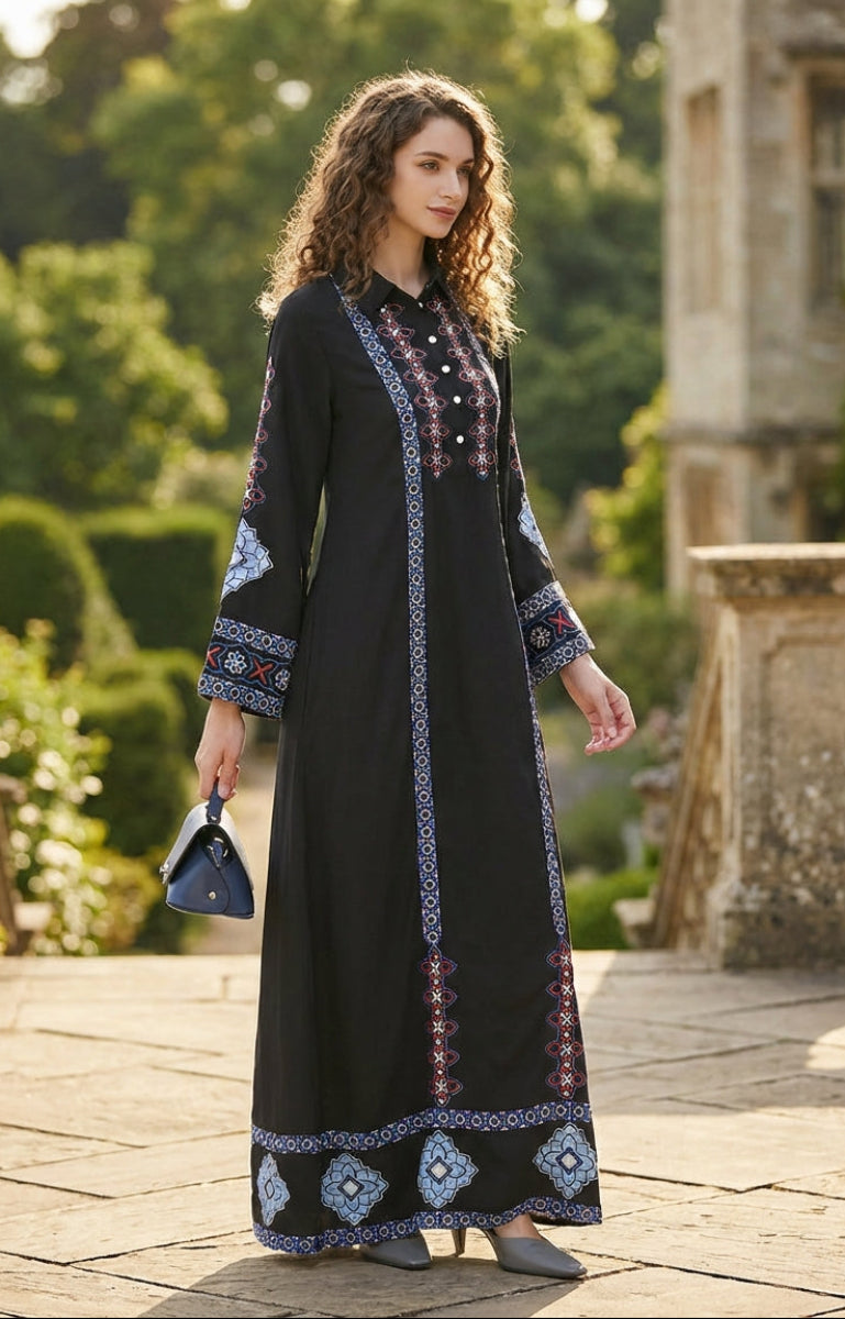 Woman in a black dress with blue embroidery standing in an outdoor setting with greenery and stone architecture.