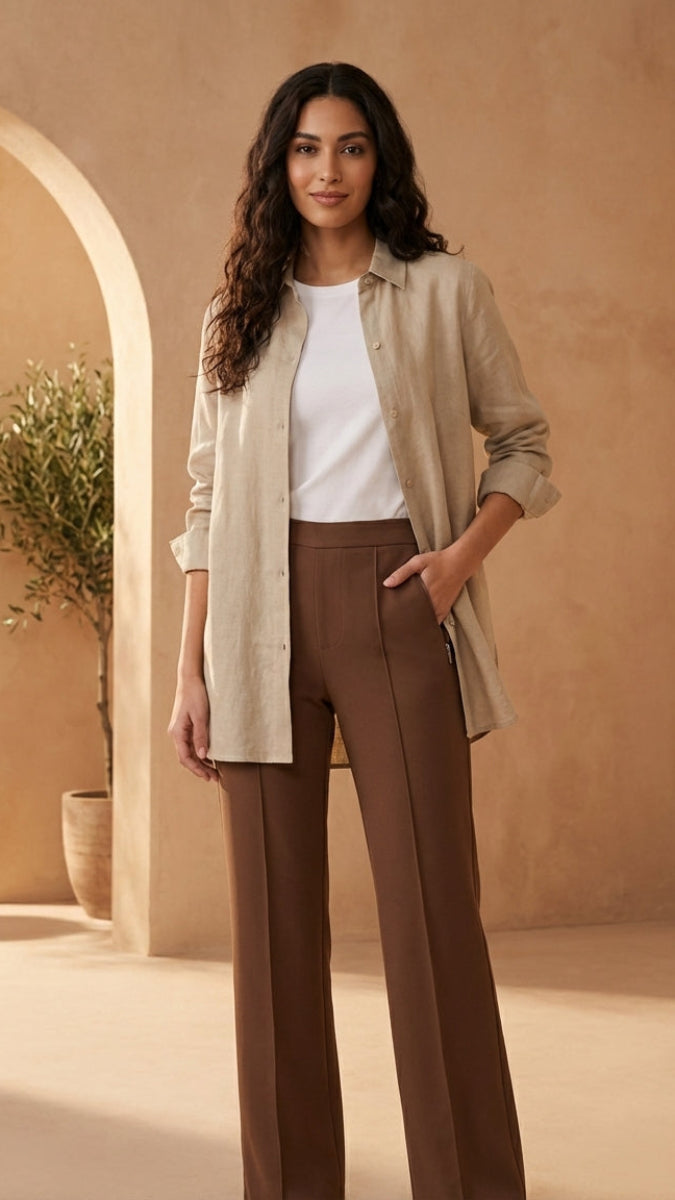 Woman in beige shirt and brown pants standing in a warm-toned architectural setting.