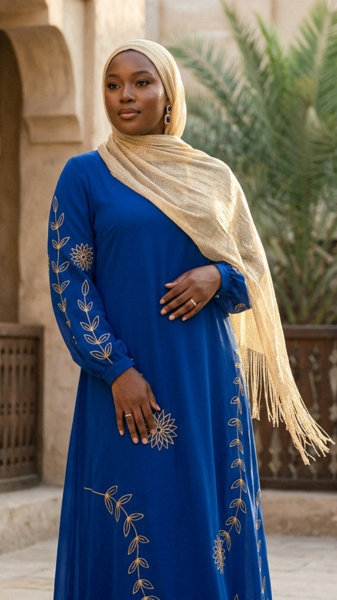 Woman in a blue dress with gold embroidery and beige hijab standing in a courtyard.