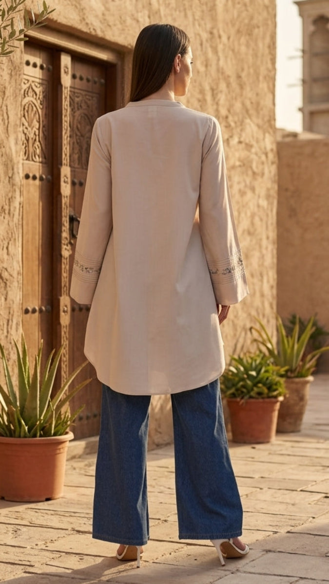 Woman in a beige coat and blue pants walking towards a traditional building with a wind tower.