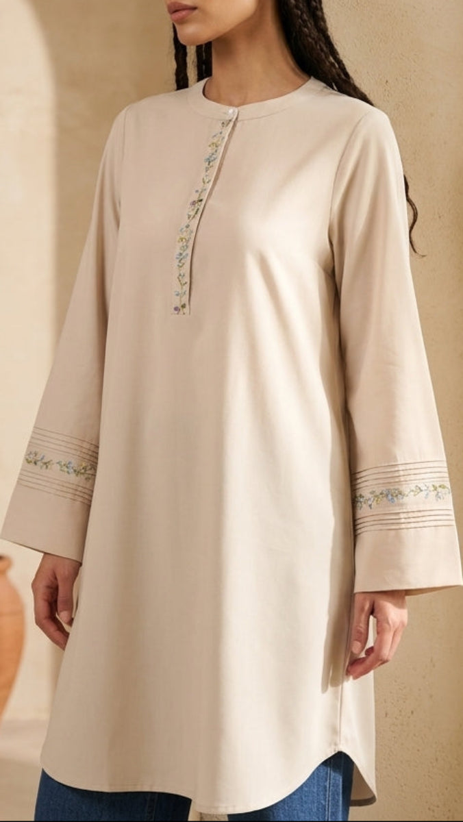 Woman wearing a beige long-sleeve top with embroidery in a warm-toned interior setting.