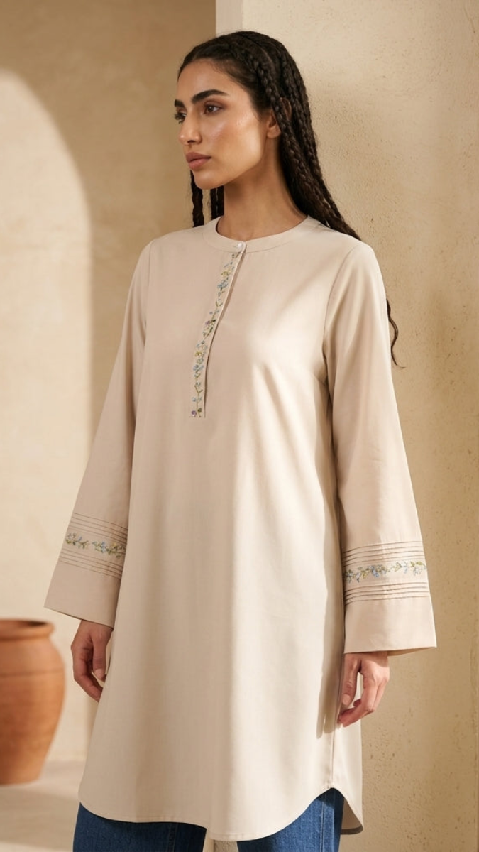 Woman wearing a beige long-sleeve top with embroidery in a warm-toned interior setting.