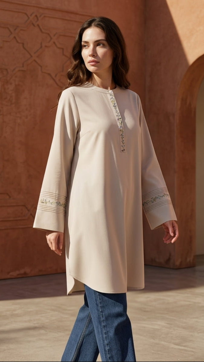 Woman wearing a long beige dress with intricate patterns in a warm-toned architectural setting.