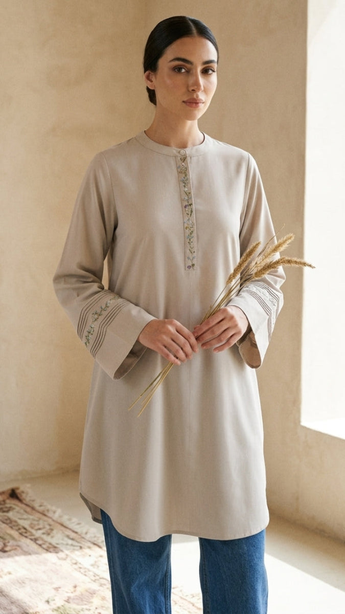 Woman wearing a beige long-sleeve top with embroidery, holding wheat stalks in a room with a textured wall and window.