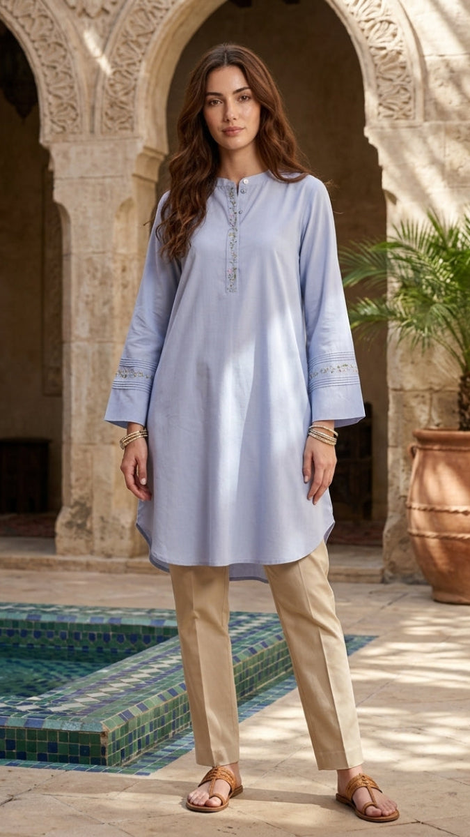 Woman in a light blue dress and beige pants standing in a sunlit courtyard with arches and a pool.