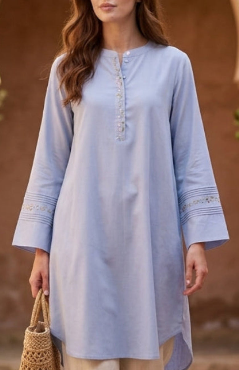 Woman in a light blue dress and beige pants walking in a sunlit courtyard with arches and potted plants.