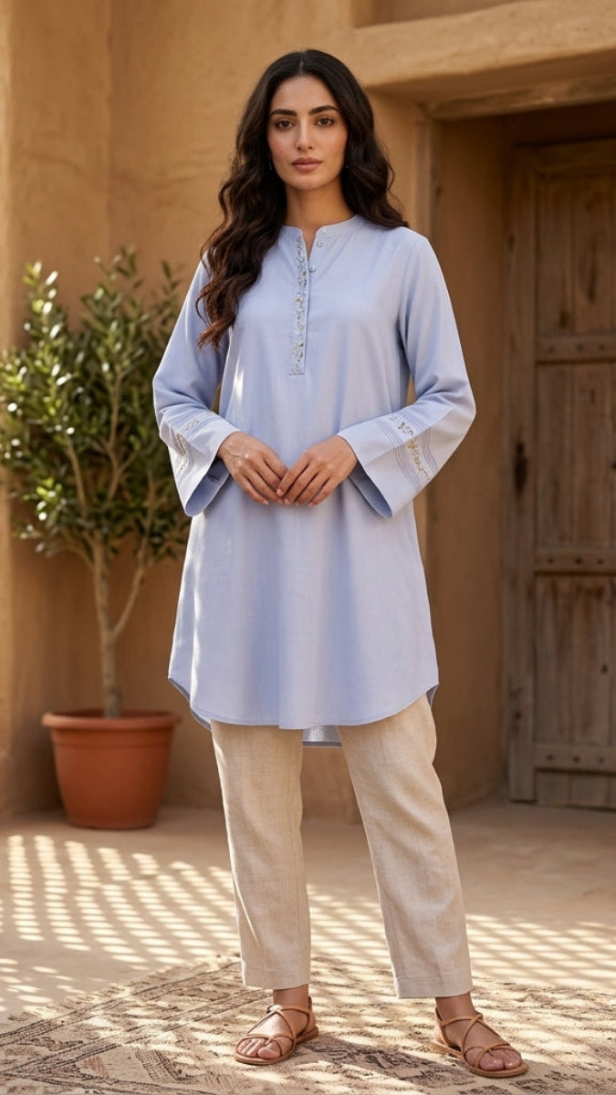 Woman in a light blue kurta and beige pants standing in a sunlit outdoor setting with plants and a wooden door.