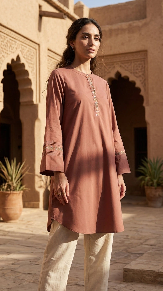 Woman wearing a long, embroidered pink dress in a sunlit courtyard with architectural elements.