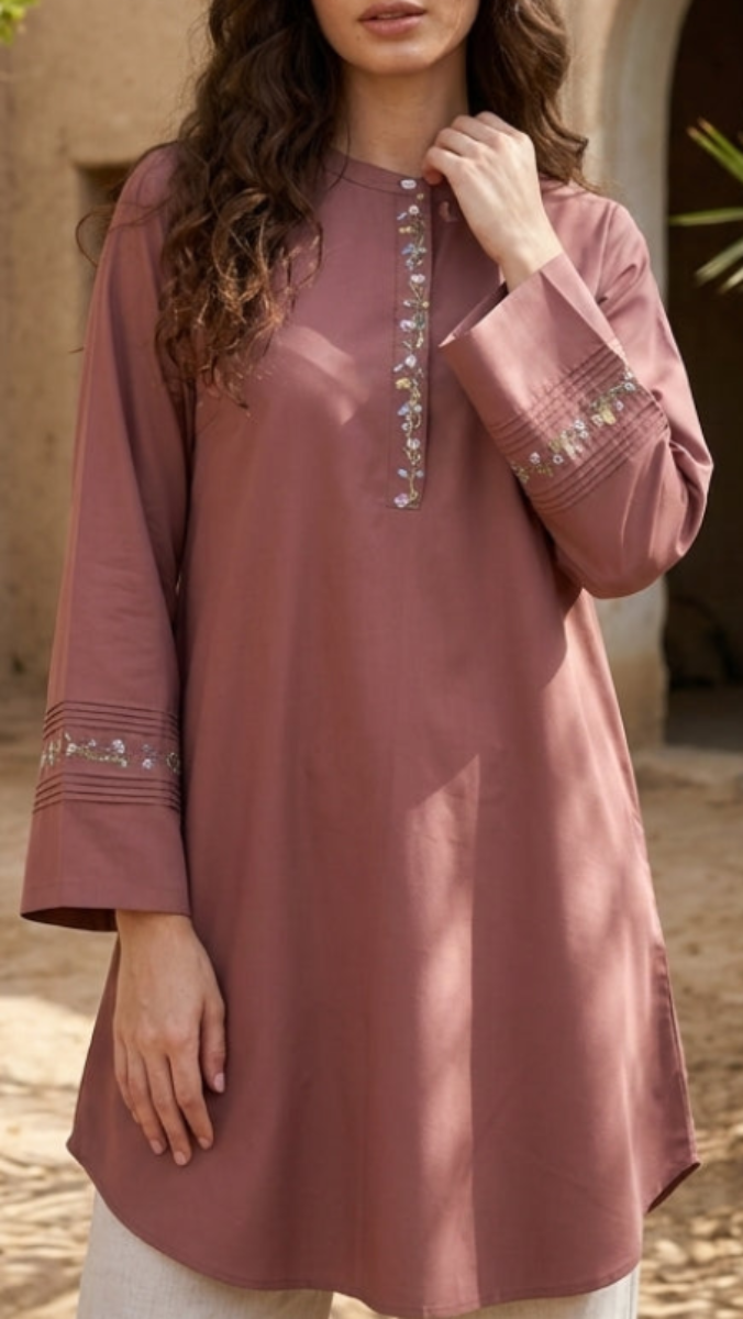 Woman wearing a mauve embroidered top and white pants standing in front of a traditional architectural background.