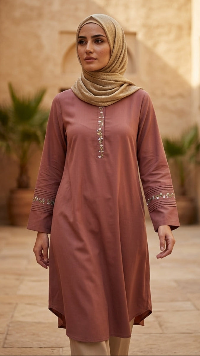 Woman in a long, light brown dress with a headscarf standing in a sunlit courtyard.