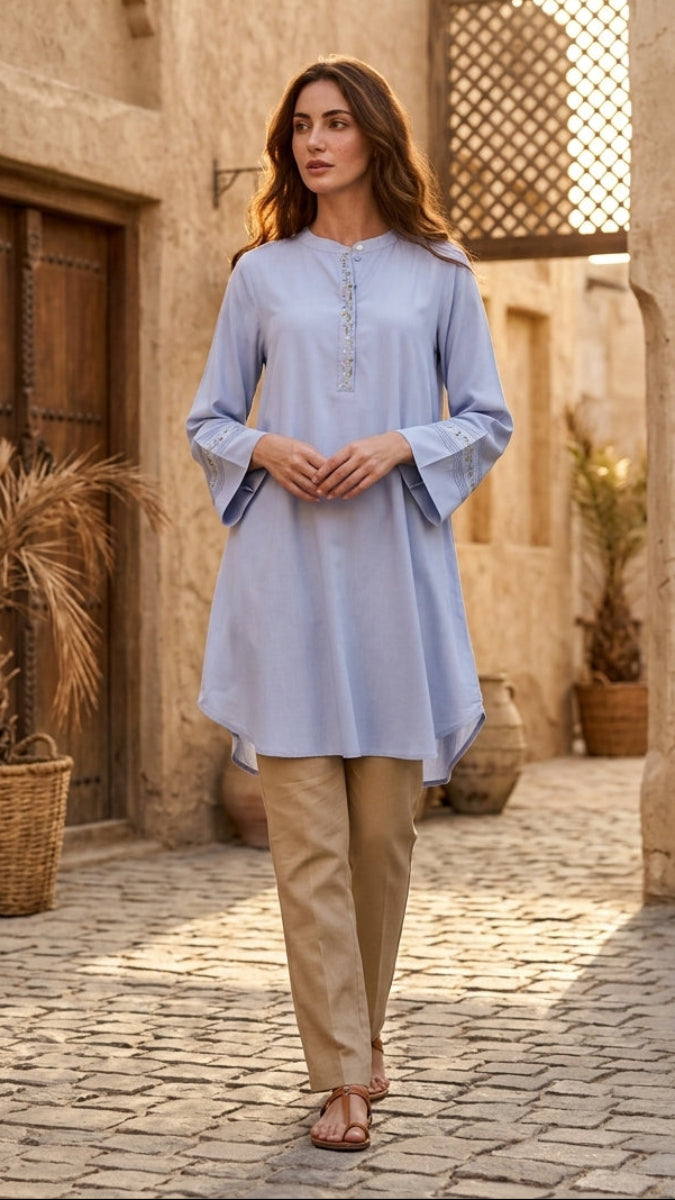 Woman in a light blue dress and beige pants walking through an arched stone passageway.