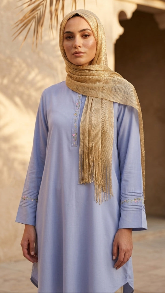 Woman wearing a light blue dress and beige hijab standing in front of a sandy architectural background.