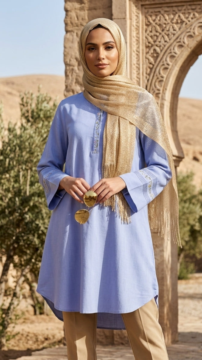 Woman in a blue dress and beige hijab standing in front of an archway with desert landscape.