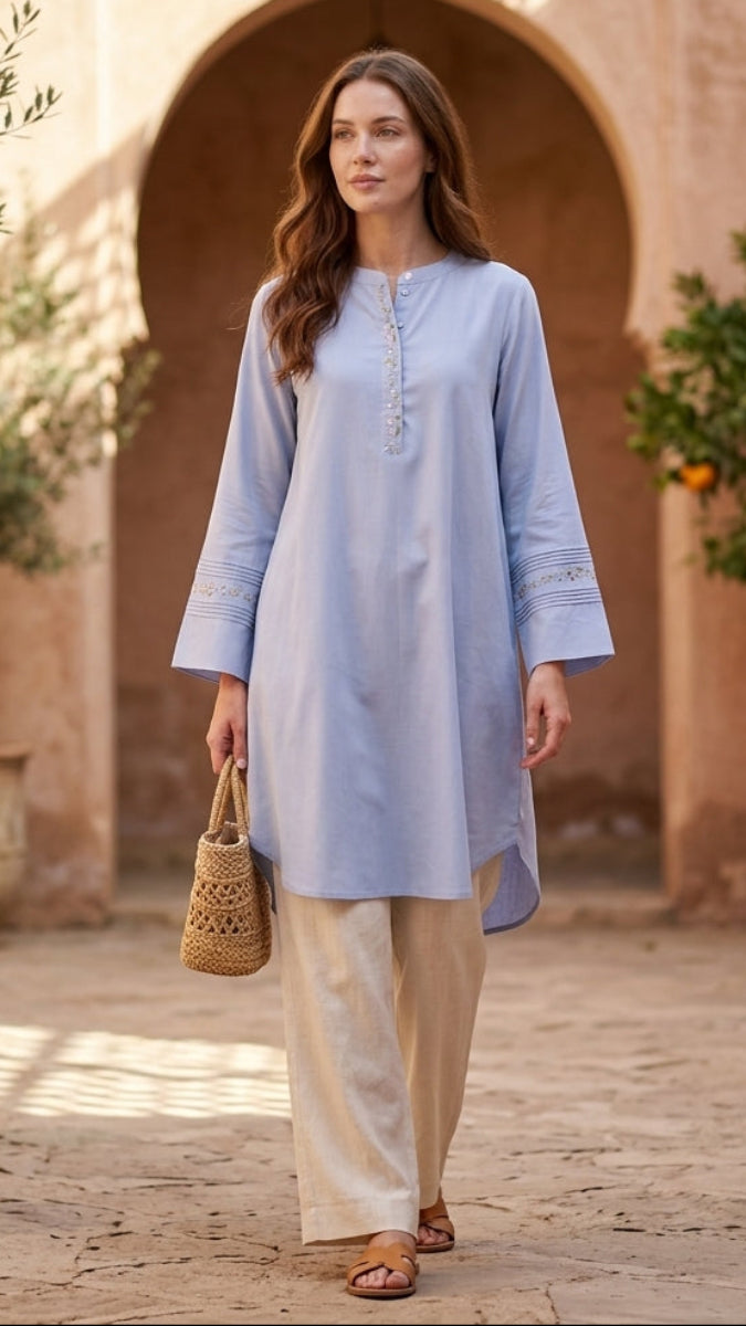 Woman in a light blue dress and beige pants walking in a sunlit courtyard with arches and potted plants.