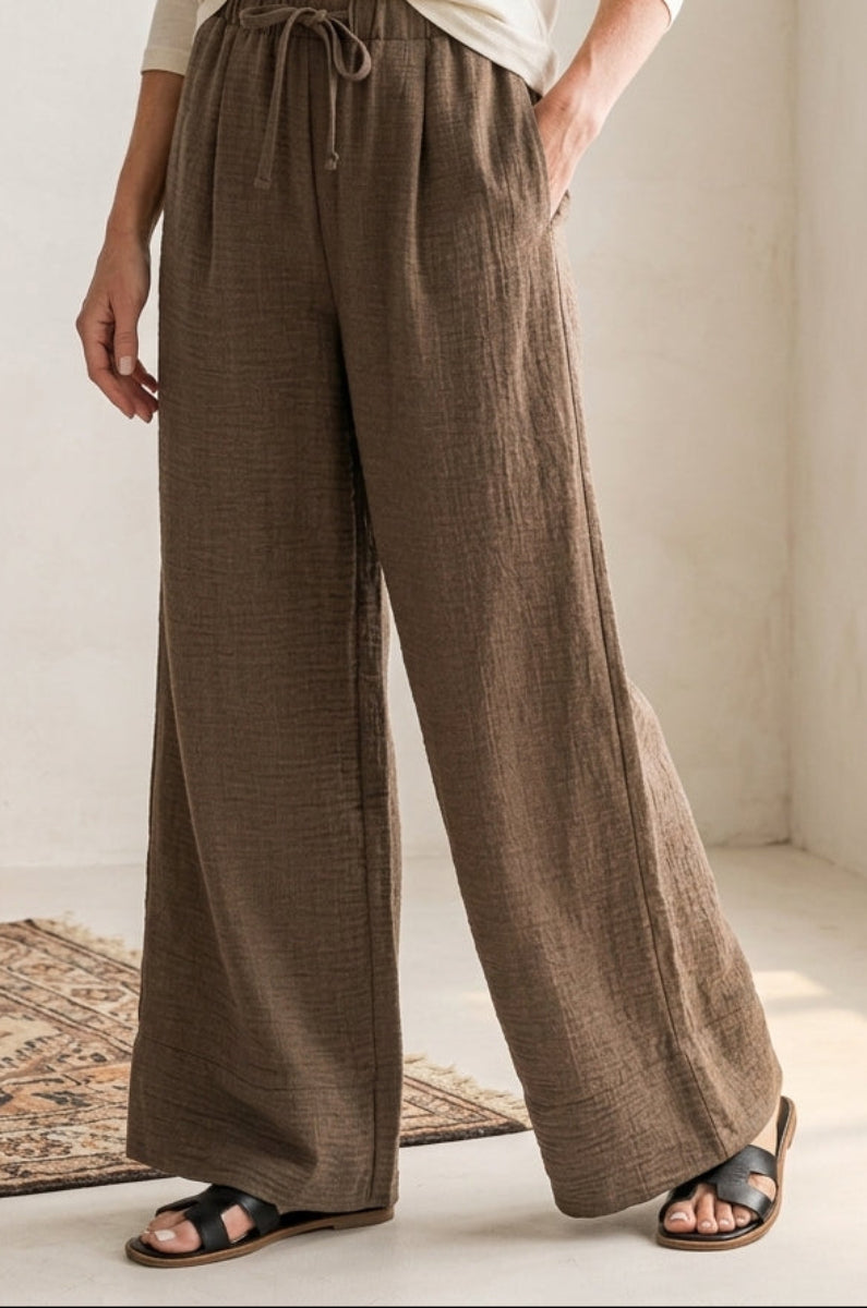 Person wearing brown wide-leg pants in a bright room with a lamp and rug.
