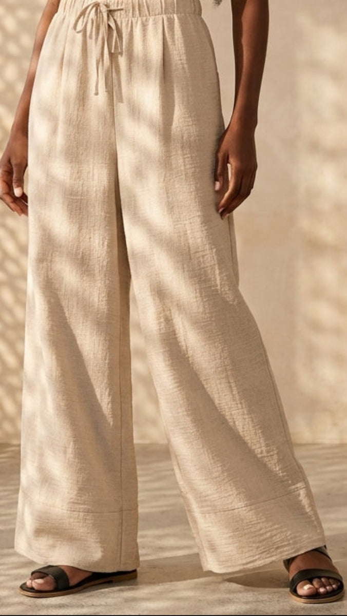 Person wearing beige wide-leg pants standing in a sunlit room with a textured wall.