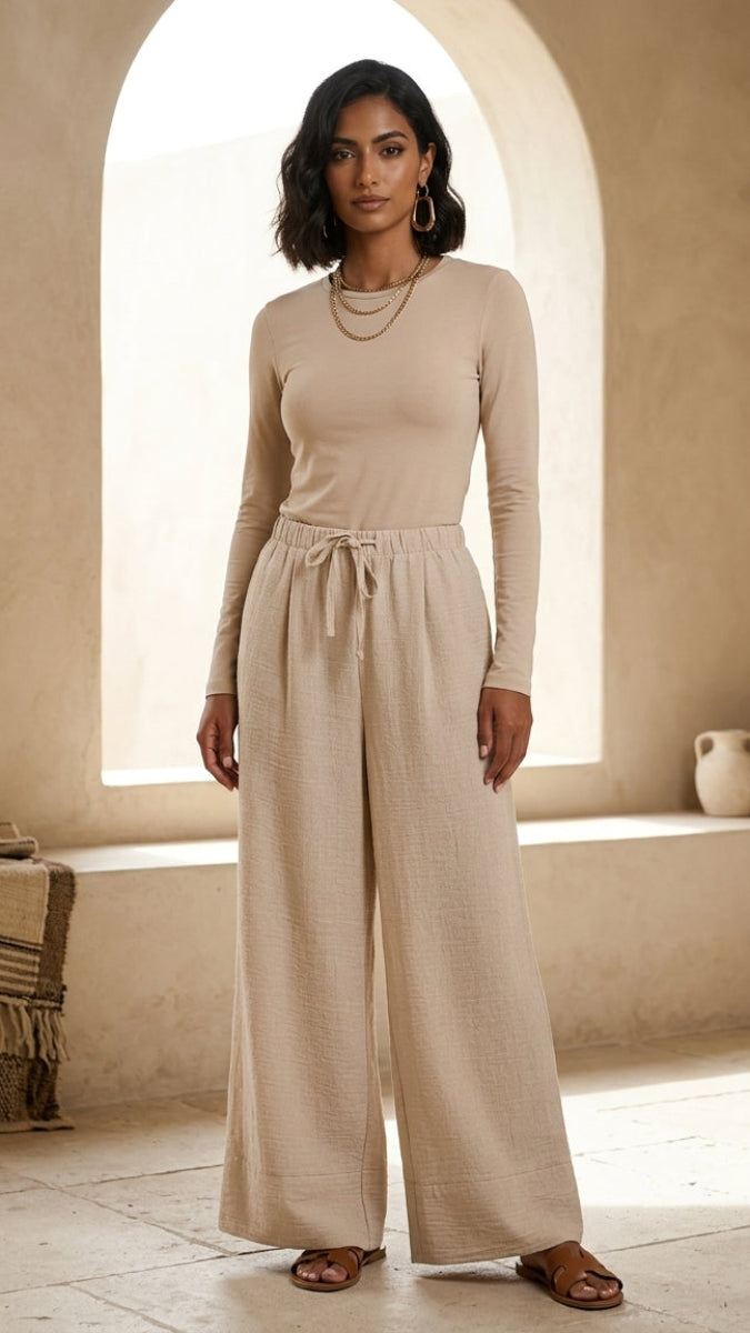 Woman wearing a beige long-sleeve top and wide-leg pants in a sunlit room.