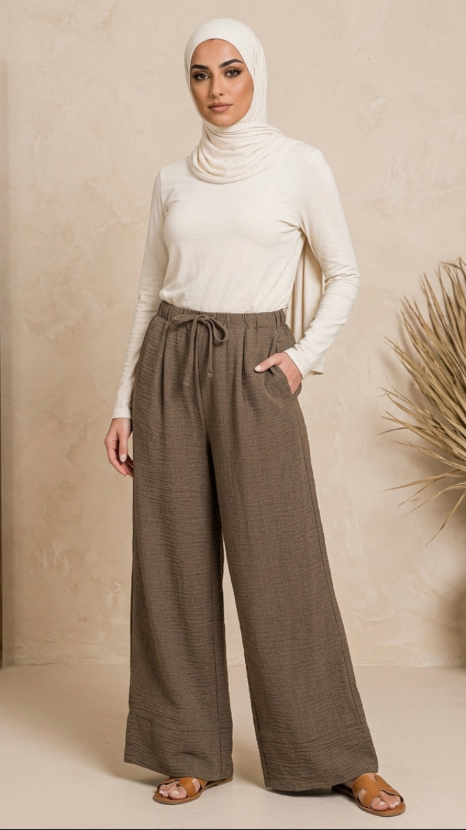 Woman wearing a hijab and white top with brown wide-leg pants against a beige wall.