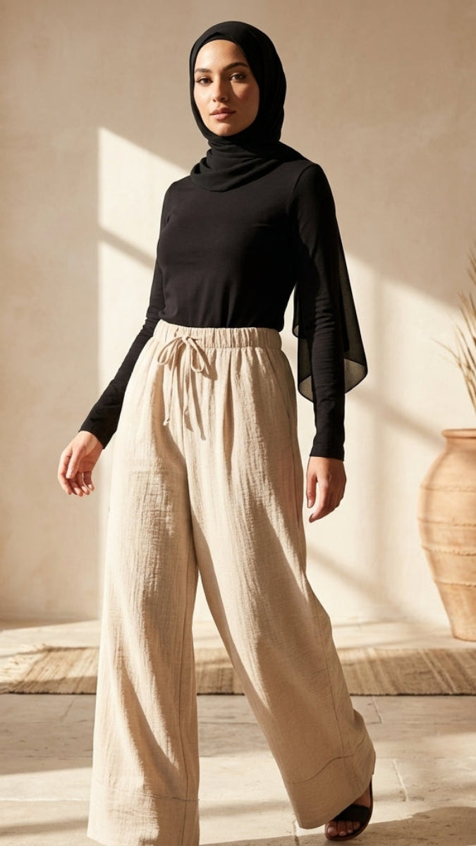 Woman wearing a black top and beige pants in a sunlit room with a vase and plant.