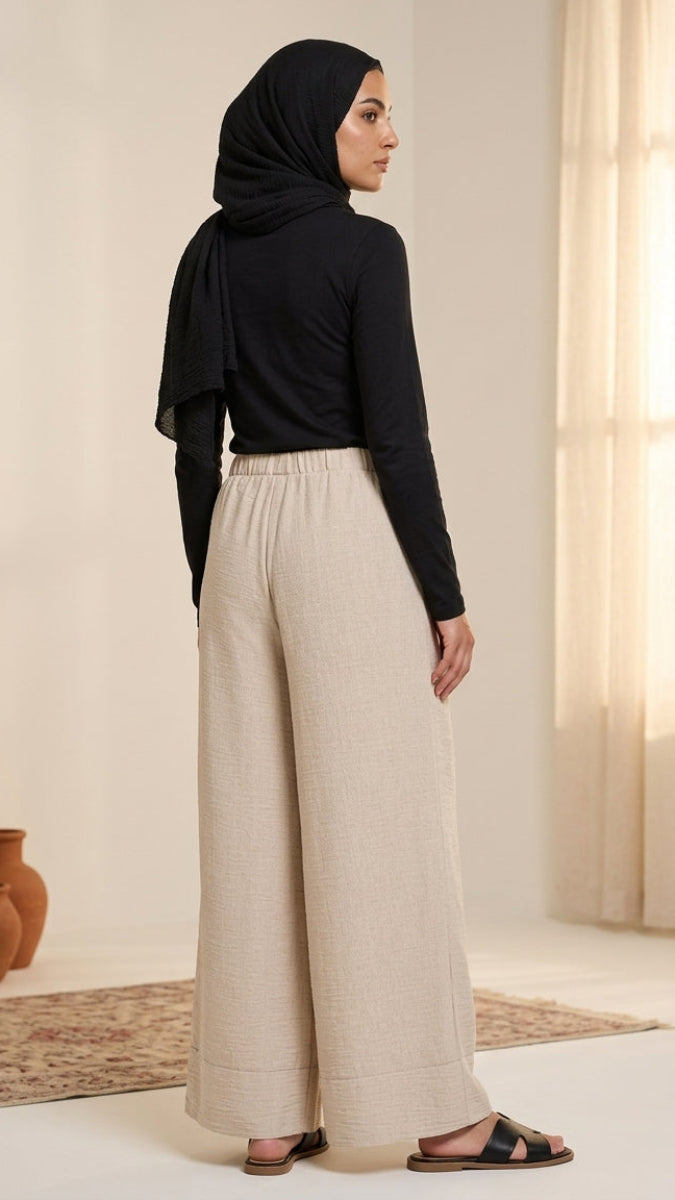 Woman wearing a black hijab and beige pants standing in a room with light-colored walls and a window.