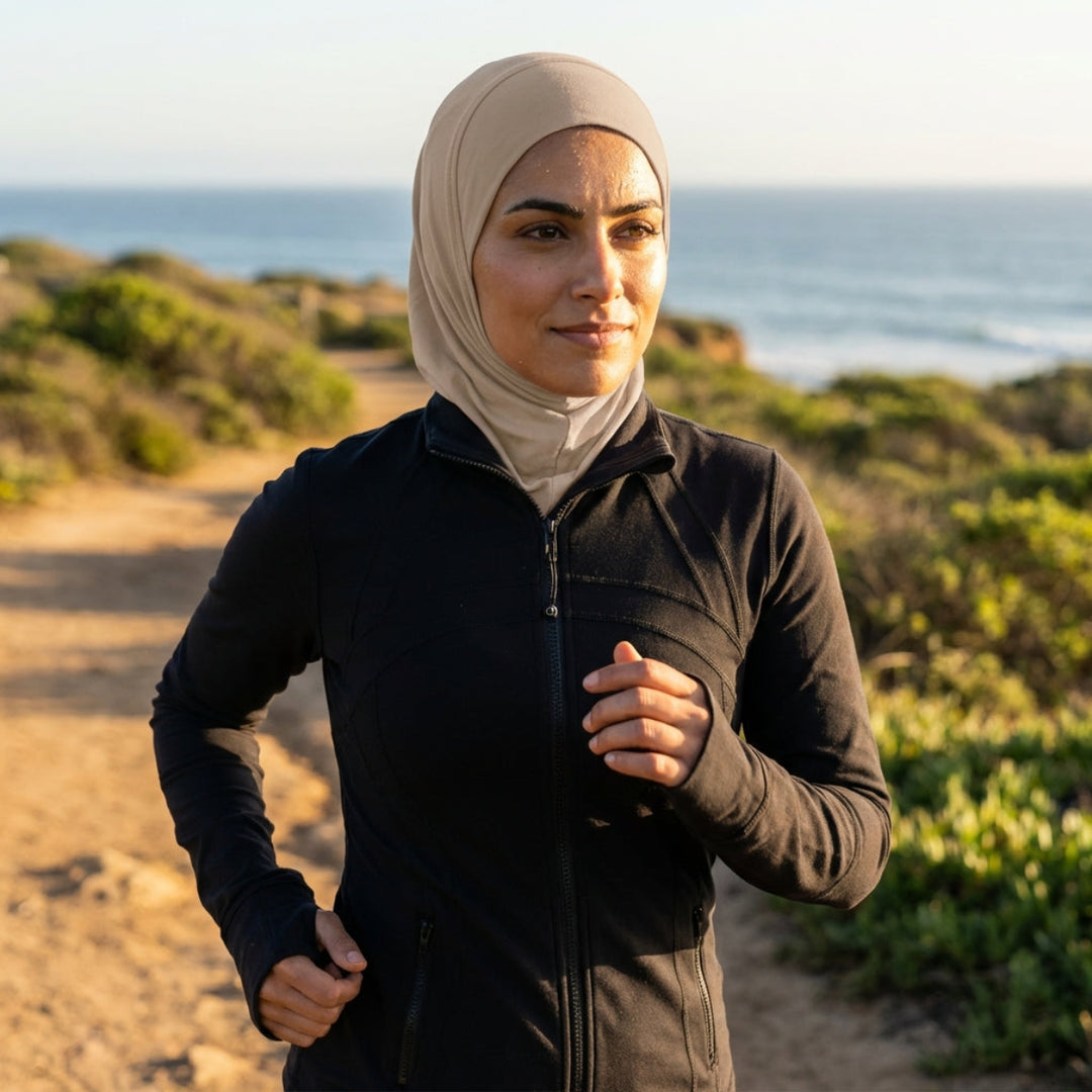 Woman in a black jacket and beige hijab standing on a trail with ocean in the background