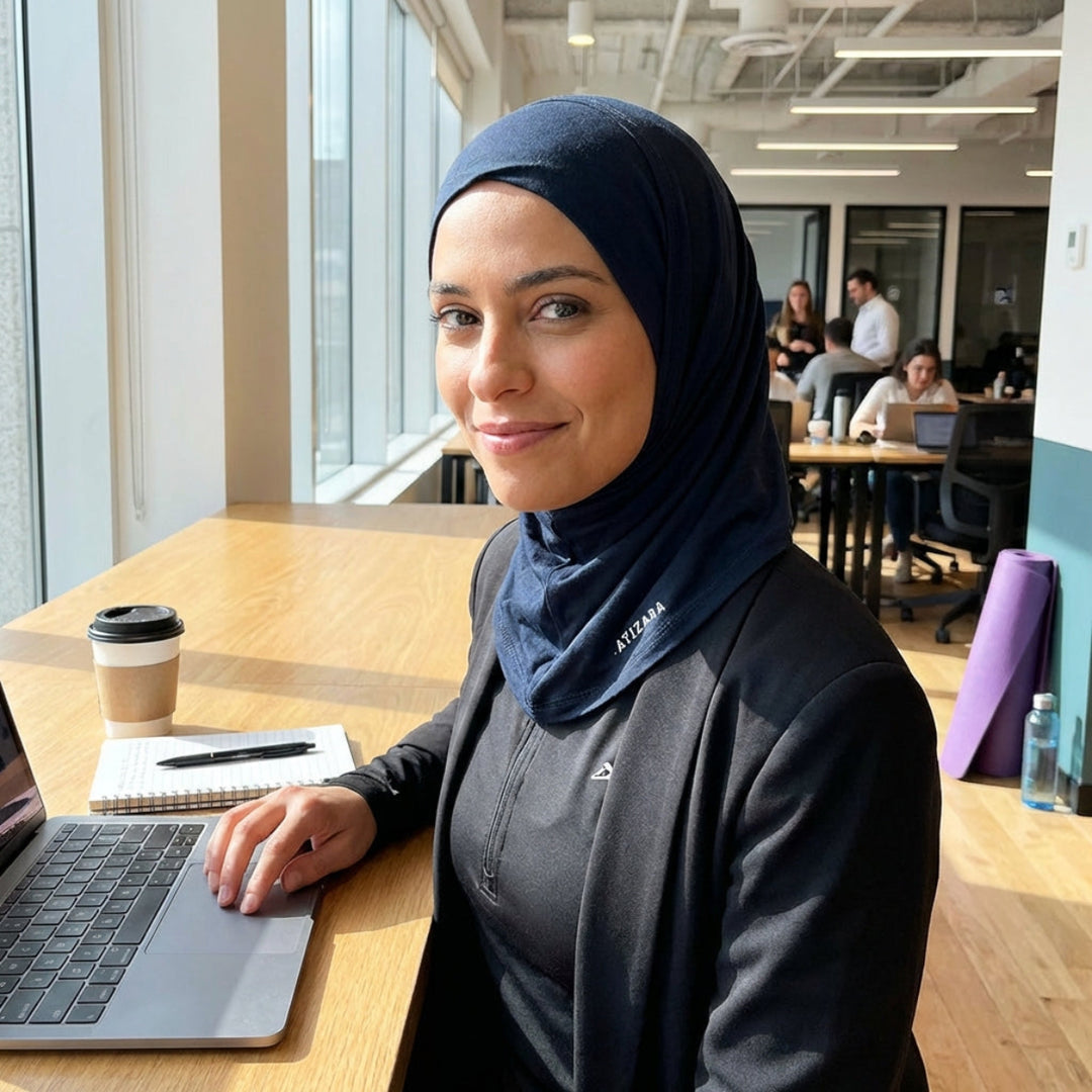 Woman wearing a hijab sitting at a desk in an office with a laptop and coffee.