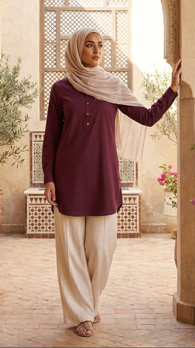 Woman in a burgundy top and beige hijab standing in an outdoor setting with arches and plants.