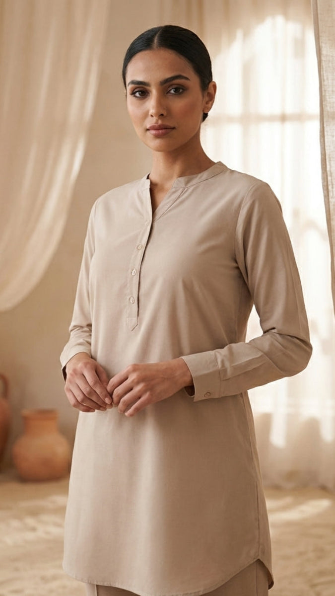 Woman wearing a beige kurta standing in a softly lit room with curtains and vases.