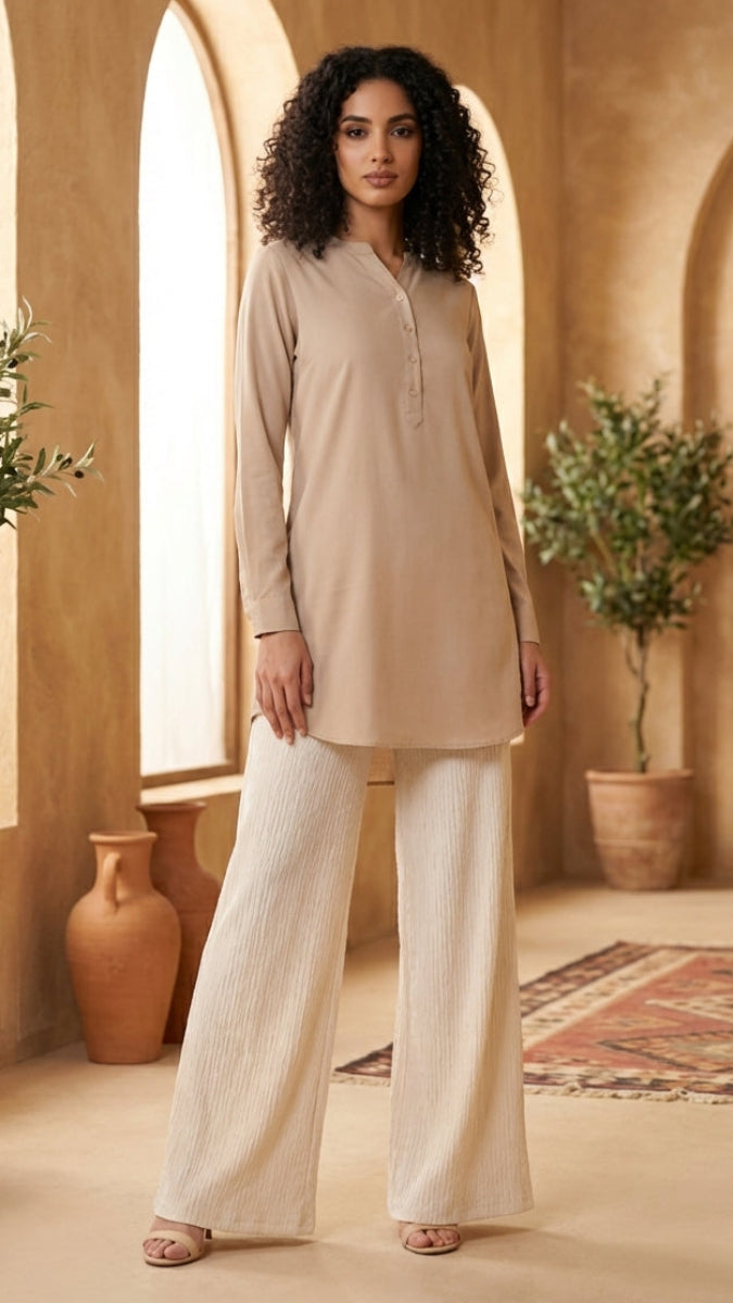 Woman in beige top and white pants standing in a warm-toned room with plants and a wooden door.