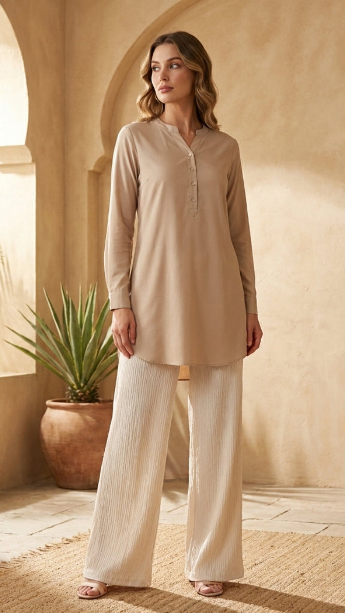 Woman in beige top and white pants standing in a sunlit room with plants.