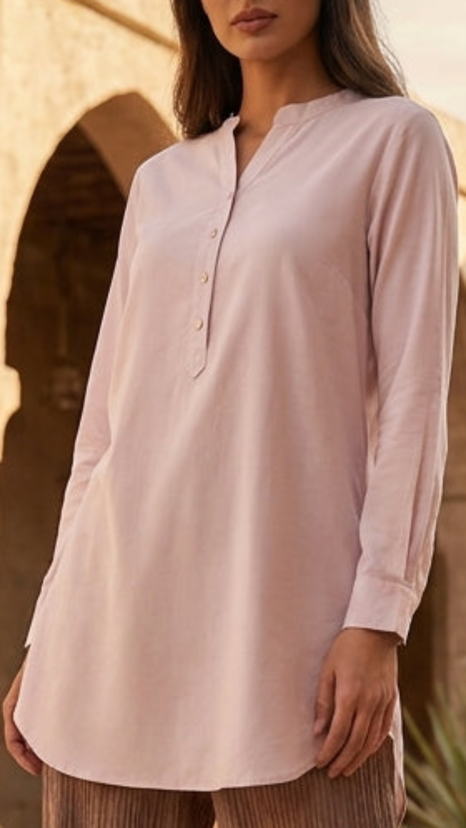 Woman in a light pink top and brown pants standing in an arched stone building with palm trees.