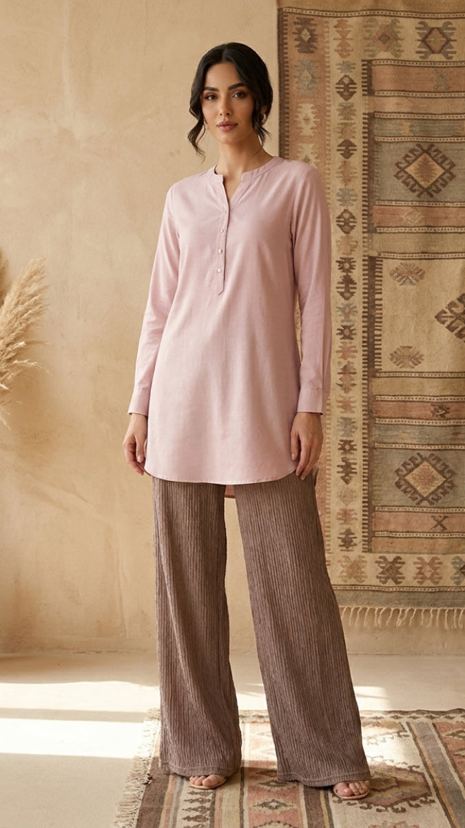 Woman wearing a pink top and brown pants standing in a room with decorative rugs and a vase.