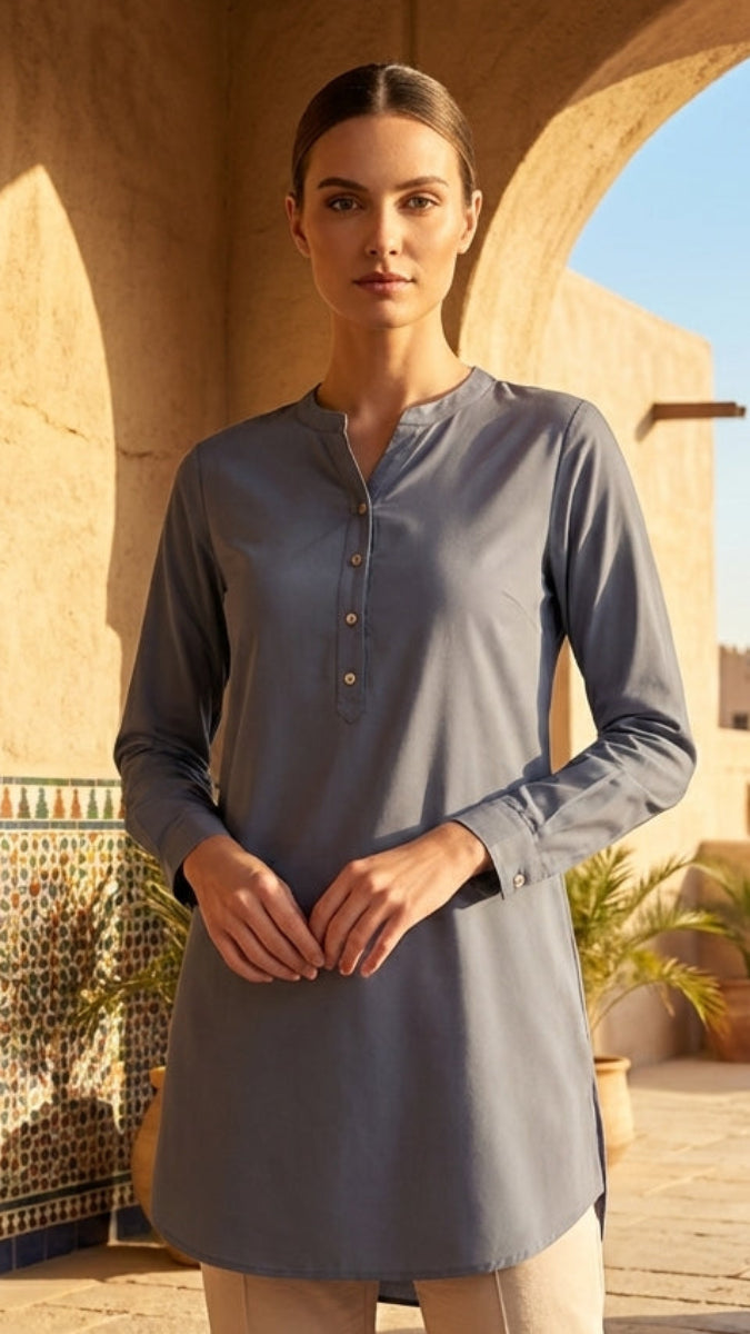 Woman in a long, light gray shirt standing in a sunlit outdoor setting with arches and plants.