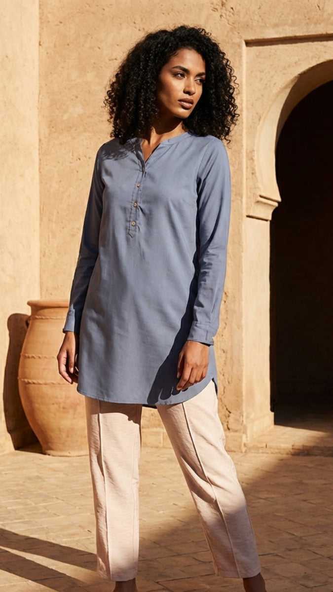 Woman wearing a blue long-sleeve shirt and beige pants standing in front of a stone building with arches.