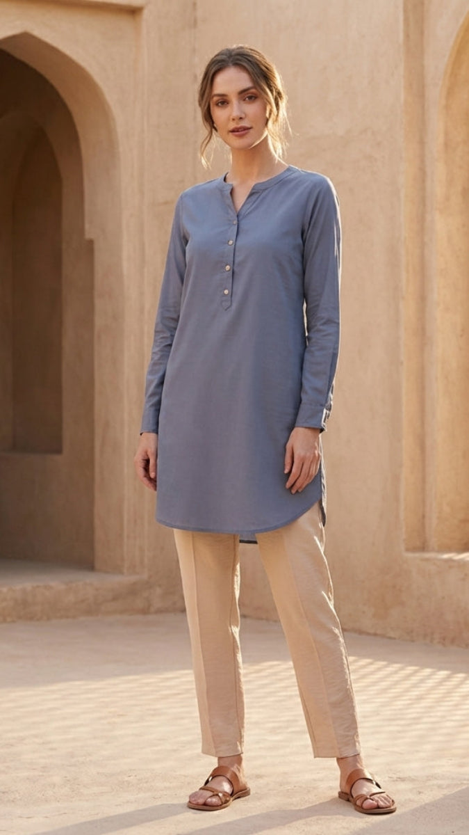 Woman wearing a blue long-sleeve shirt and beige pants standing in a sunlit architectural setting.
