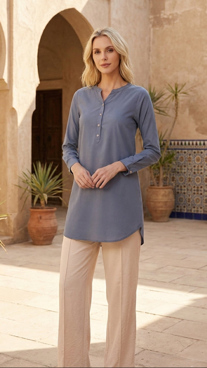 Woman wearing a blue shirt and beige pants standing in a sunlit courtyard with arches and potted plants.