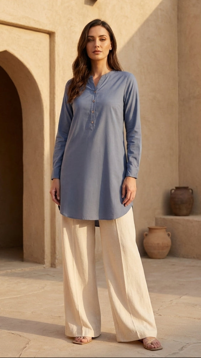 Woman wearing a blue top and beige pants standing in front of a beige wall with an archway.