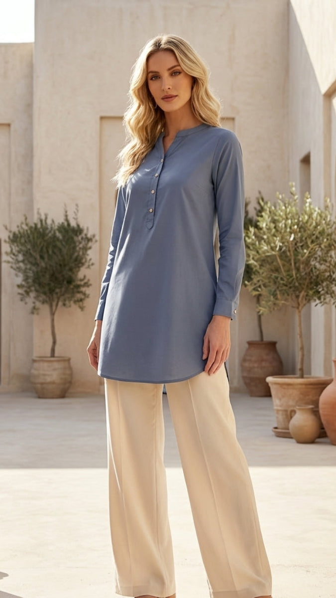 Woman wearing a blue long-sleeve top and beige pants in an outdoor setting with architectural elements and plants.