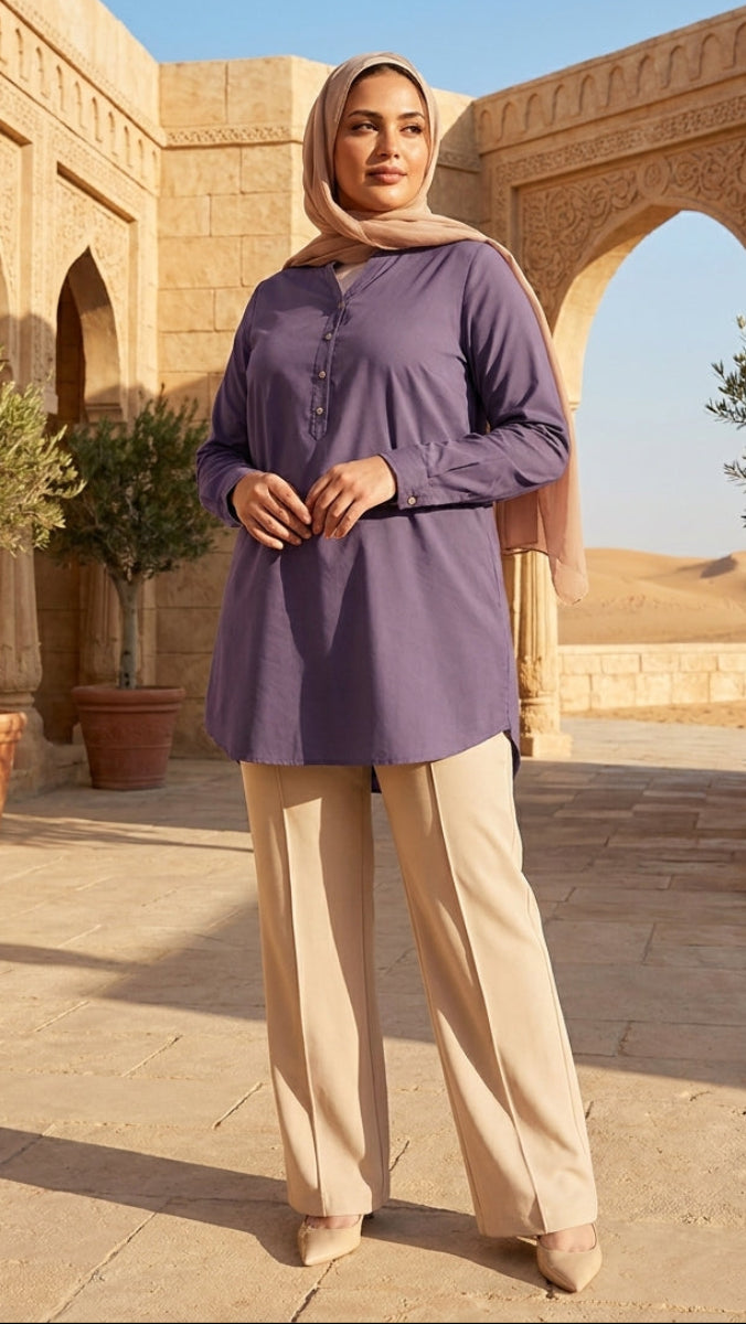 Woman in a purple shirt and beige hijab standing in front of architectural structures with plants.