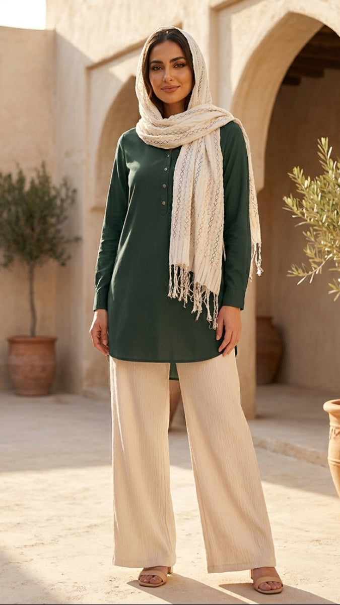 Woman in green top and beige scarf standing in a sunlit courtyard with arches and potted plants.