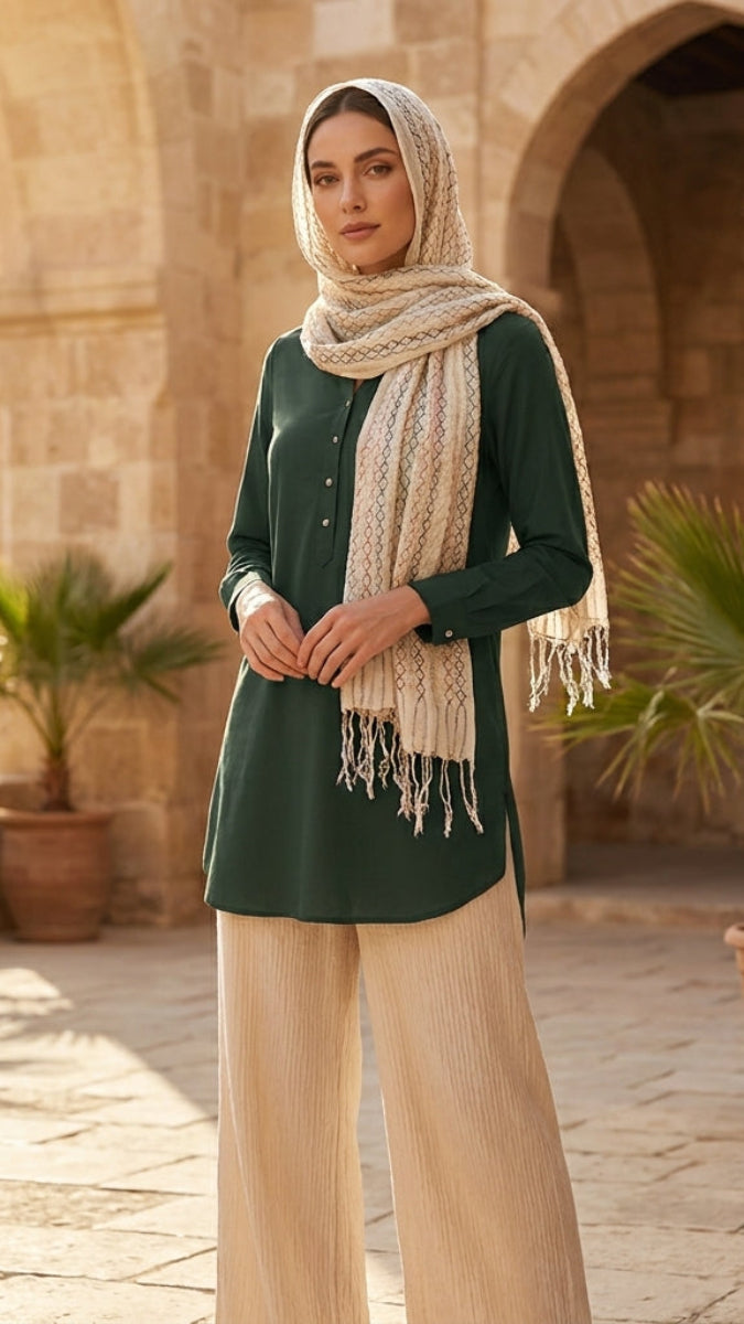 Woman in a green top and beige scarf standing in front of stone arches and potted plants.