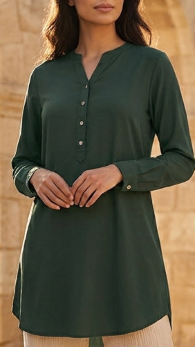 Woman in a green top and beige pants standing in front of stone arches and potted plants.