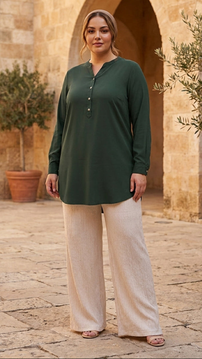 Woman wearing a green long-sleeve top and beige pants standing in a sunlit courtyard with arches and potted plants.