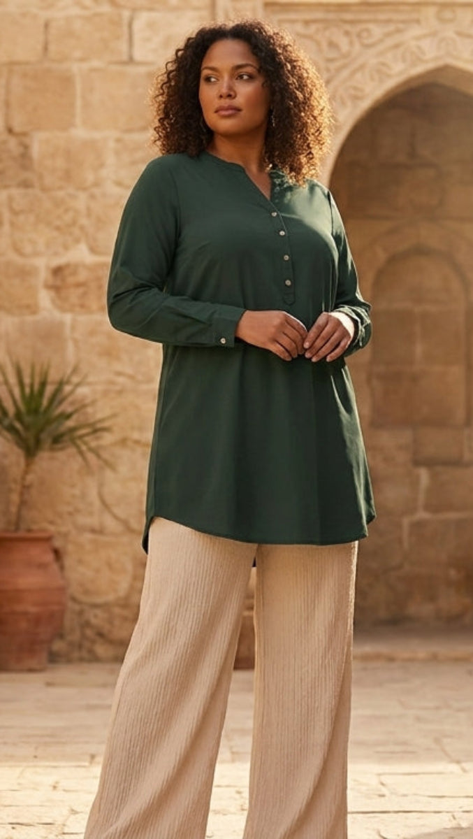 Woman in a green shirt and beige pants standing in a sunlit stone courtyard with potted plants.