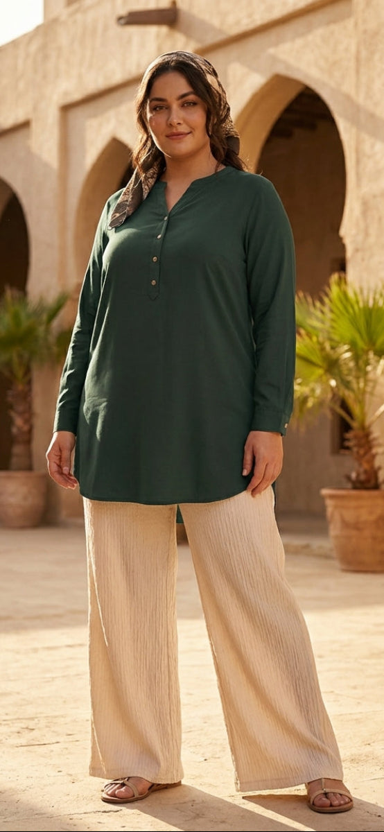 Woman wearing a green long-sleeve top and beige pants standing in front of arched stone buildings with palm trees.