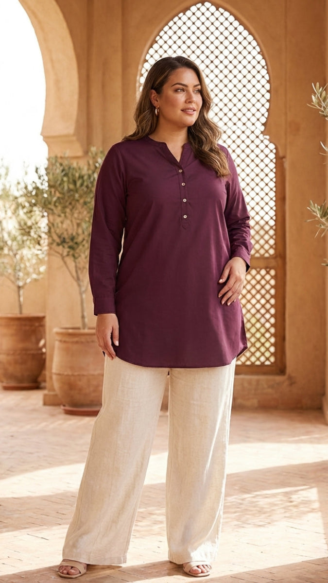Woman wearing a purple shirt and white pants standing in a sunlit outdoor setting with arches and plants.